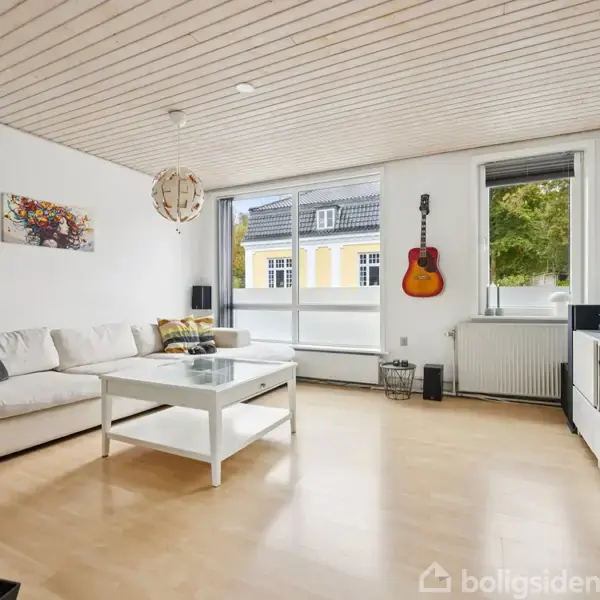 Stue med lys trælaminatgulv, en stor hvid sofa og et hvidt sofabord. Guitaren hænger ved vinduet, og der er grønne planter. Vinduerne viser gule bygninger udenfor.