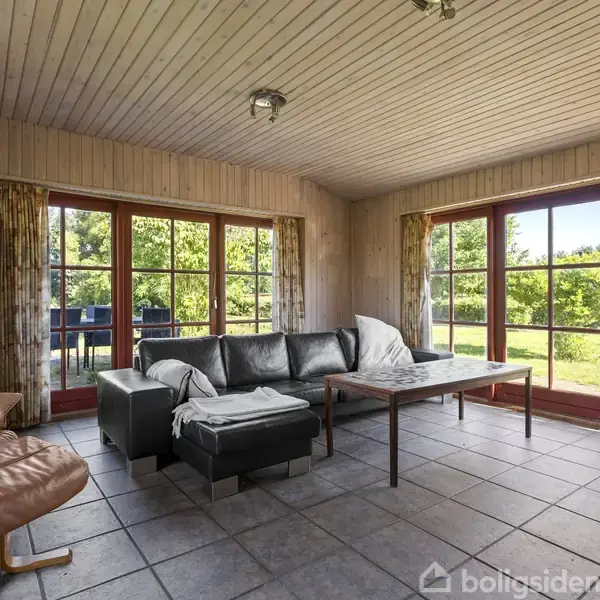En stue med en sort lædersofa står foran store vinduer, der giver udsigt til en grøn have. En lænestol er til venstre, og et sofabord står foran sofaen.