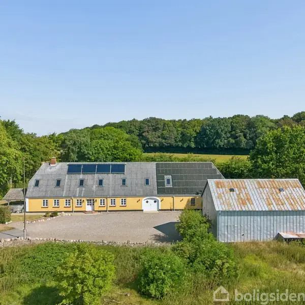 Gult hus med solpaneler på taget står ved siden af en stor grå lade. Omgivet af marker og træer under en blå himmel. En bil er parkeret ved huset.