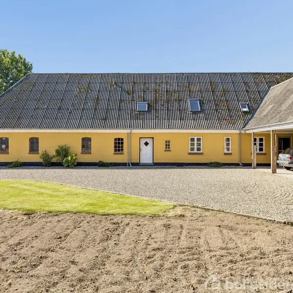 Gul landbygning med mørkt tag og flere små vinduer. Står i et åbent landligt område med grønne marker og grusbelagt gårdsplads. En bil er delvist synlig i en garage.