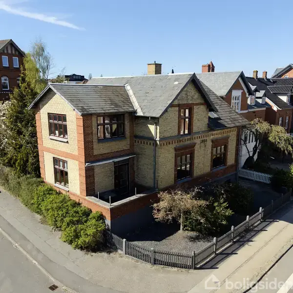 Et to-etagers hus, lavet af rød- og gulmursten, står i et villakvarter med flere lignende bygninger omkring. En hvid bil er parkeret langs vejen ved siden af huset.
