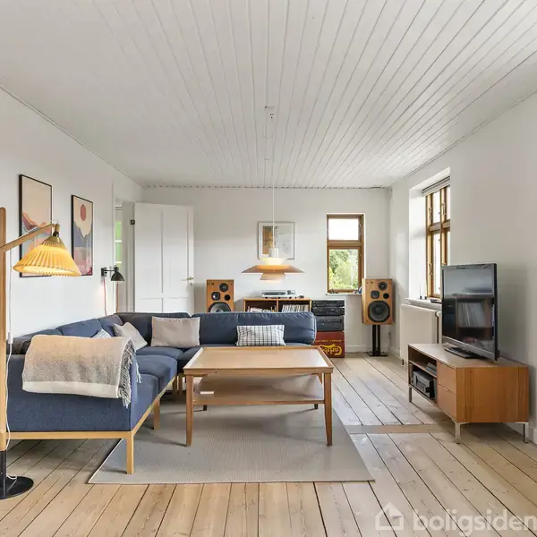 Et hvidt hus med sort tag står i en boligbydel. Huset har flere vinduer og en indkørsel. Det er omgivet af grønne buske, træer og andre hjem i baggrunden.