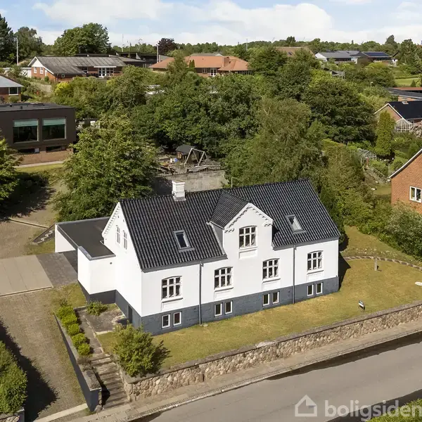Et hvidt hus med sort tag står på en stenbelagt forhøjning. Det er omgivet af en græsplæne og et fortov i et beboelsesområde med andre huse.