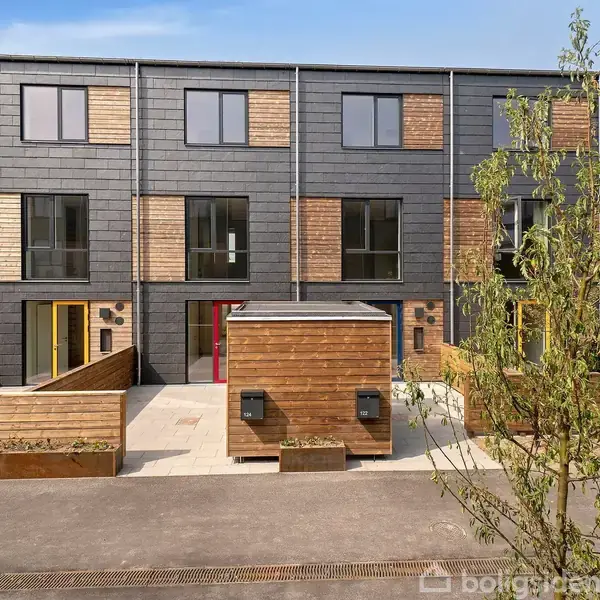 Rækkehus med mørk facade og trædetaljer står i en moderne boligklynge. Foran huset er en indgang med små træskure og en flisebelagt gårdsplads, omgivet af et enkelt træ.