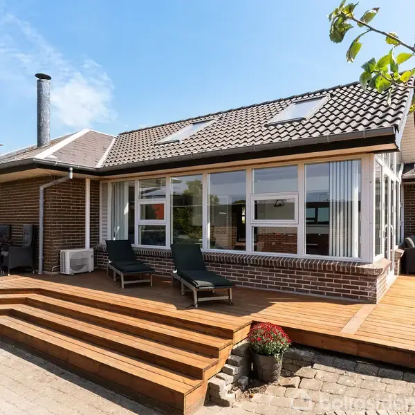 Murstenshus med store vinduer og brændeovn står på en terrasse med trætrapper. To liggestole står foran huset. Omkring er der blå himmel og en frugtplante med æbler.