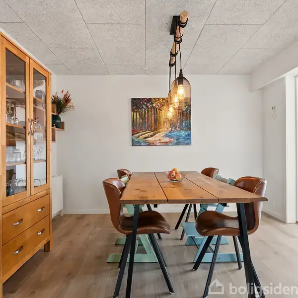 Et spisebord med seks stole står i et lyst rum. En trævitrine indeholder glas. En farverig maleri hænger på væggen, og en lampeskærm hænger over bordet.