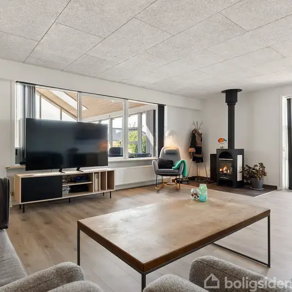 Stue med grå sofaer omkring et træbord. Et tv står på et skab, mens en brændeovn giver varme. Store vinduer og glasdøre lukker masser af lys ind. En sort vægur hænger på væggen.