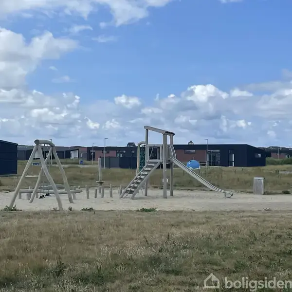 Legeplads med gyngestativ og rutsjebane placeret i et åbent græsareal omgivet af moderne huse. En bænk og et affaldsspand står ved siden af legeredskaberne.