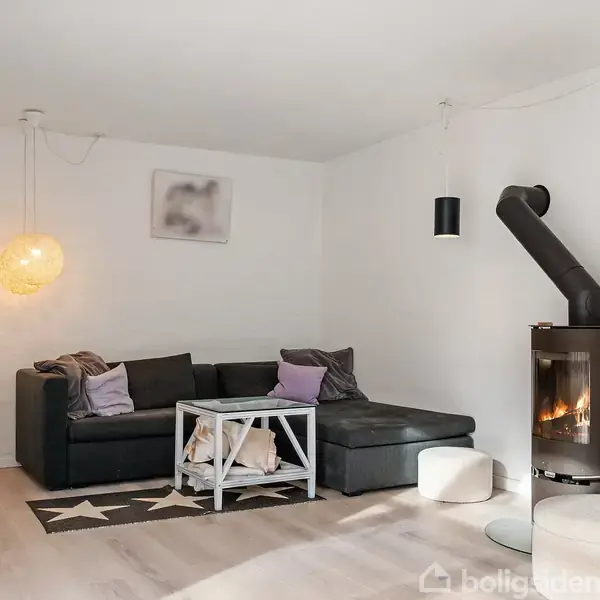 Sort sofa står i hjørnet af en stue ved siden af en brændeovn. Et sofabord med tæppe ovenpå står foran. En rund lampe hænger fra loftet, skaber en hyggelig stemning.