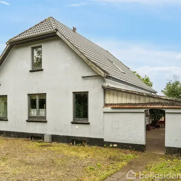 Et hvidt hus med rødt tag står stille på en grusbelagt grund. Ruderne er firkantede, og der er en tilstødende lille bygning. Træer omgiver ejendommen under en blå himmel.