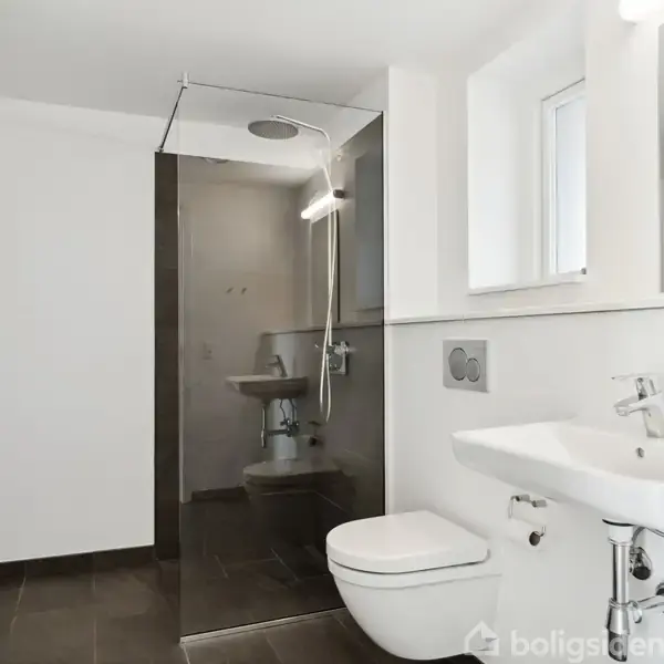 Et moderne badeværelse med en væghængt toilet og håndvask, en glasafskærmet brusekabine under et stort loftsbruserhoved. Rummet har lyse vægge, mørkt flisegulv og et lille vindue.