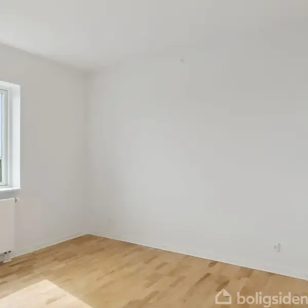 Et tomt værelse med trægulv og en lys væg. Sollys skinner gennem et vindue, der viser en grøn udsigt udenfor. Radiator under vinduet.