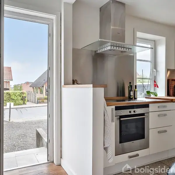 En moderne køkkenindretning med hvide skabe og træbordplader. Komfur med emhætte er i centrum. En åben dør fører til en solrig gårdsplads med huse i baggrunden.