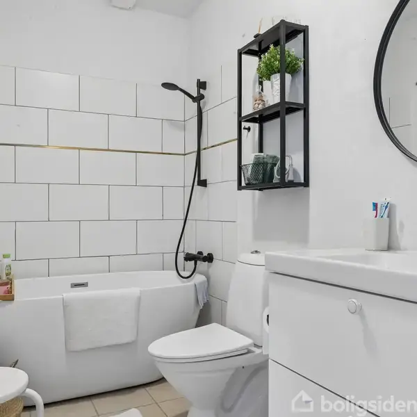 Et badeværelse med et fritstående badekar ved en hvid flisevæg. Ved siden af står toilettet, og en vask med sorte armaturer. Hylder med planter og andre genstande hænger på væggen.