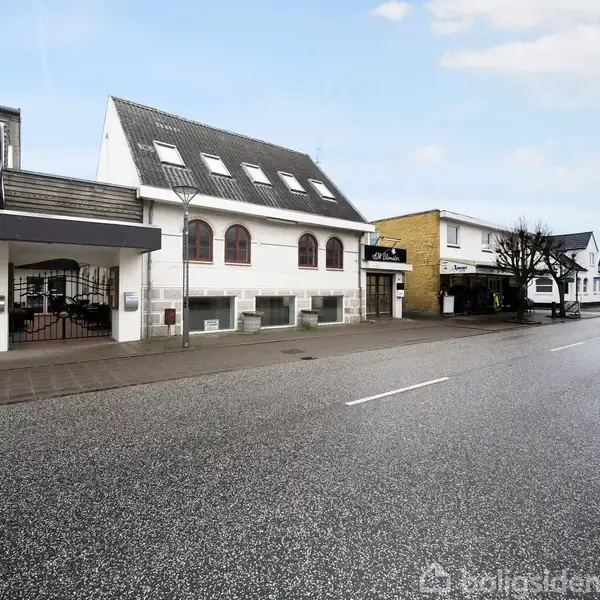 En række forskellige bygninger står stille langs en vej i et bymiljø. Bygningerne har varierende arkitektoniske stilarter, og der er få træer langs fortovet.