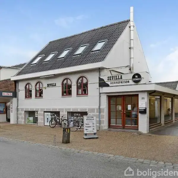 En hvid bygning med skiltene “Sevilla” og “Herrefrisør” står ved en gade. To cykler er placeret foran bygningen. Konteksten er en handelsgade med andre butikker og en biograf.