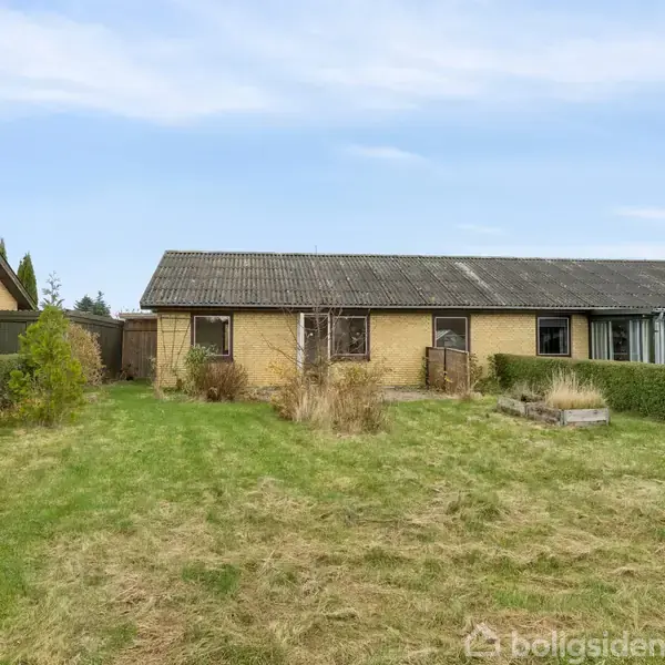 En lang, gul murstensbygning står stille i en have med græs og buske, omgivet af en lav hæk. Der er en blå himmel i baggrunden.