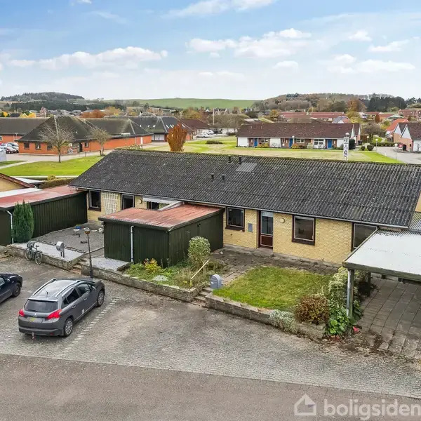 En enkelthus med græsplæne foran, flere parkerede biler, og en carport ved siden. Det ligger i et roligt boligområde med lignende huse i baggrunden og klare blå skyer på himlen.
