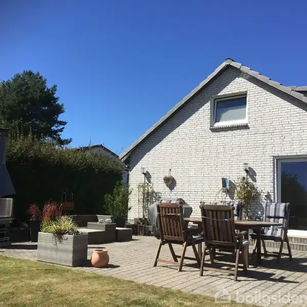 Et hvidt murstenshus med skråt tag, omgivet af en terrasse med havemøbler og planter. I baggrunden er der grønne buske og himlen er klar og blå.