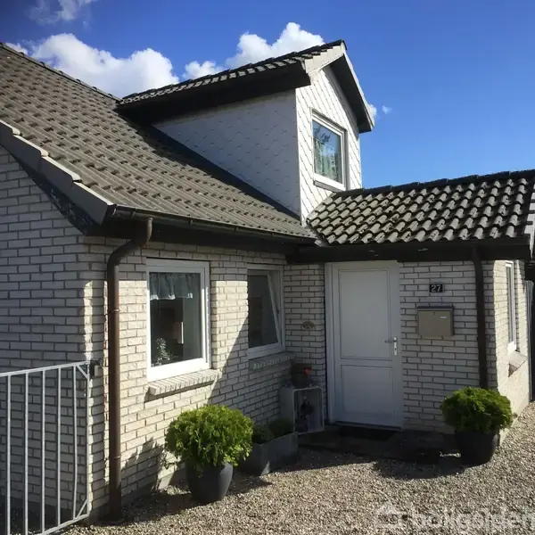 Et hvidt murstenshus med mørkt tag står i sollys. Hoveddøren er flankeret af vinduer og planter i potter. En sti af grus fører til døren. Nummeret "29" ses tydeligt.