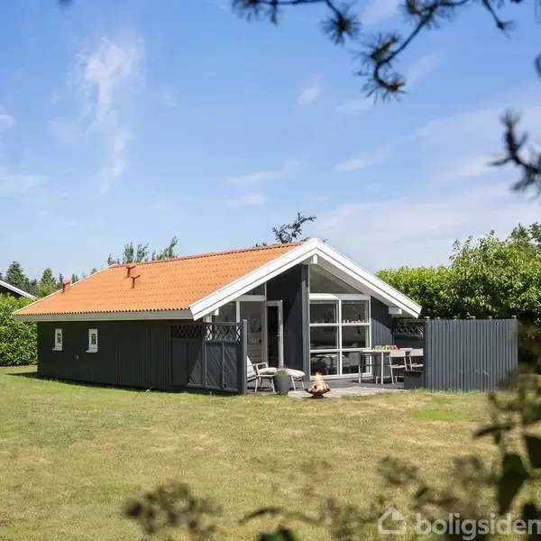 Et sommerhus med orange tegltag og mørke vægge står i en have. Der er havemøbler på terrassen. Omgivet af grønne buske og en klar blå himmel.