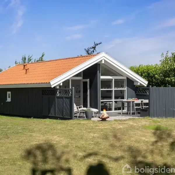 Hus med orange tag ligger i en grøn have, omgivet af buske. På terrassen står et havebord og stole. Klart solrigt vejr giver en hyggelig sommerstemning.