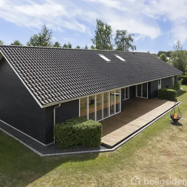 Et moderne, sort træhus med store vinduer står stille på en grøn græsplæne. Foran huset er en træterrasse. Haven er omkranset af træer og buske.