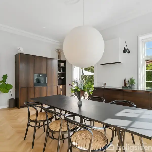 Spisebord med seks stole står i et lyst køkken-alrum. En stor kuglelampe hænger over bordet. Brune træskabe står til venstre, og store vinduer lader lys falde ind.