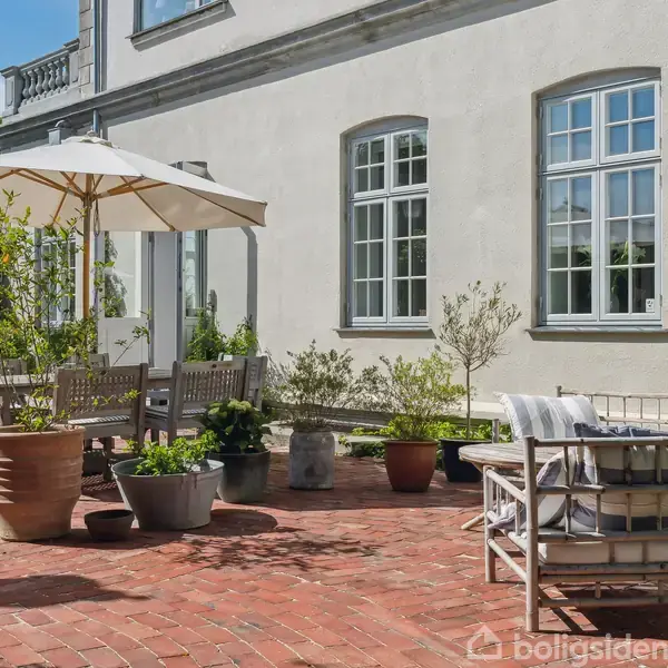En udendørs terrasse har træmøbler og en parasol, omgivet af potteplanter på røde murstensfliser. I baggrunden ses en lys facade med store vinduer.