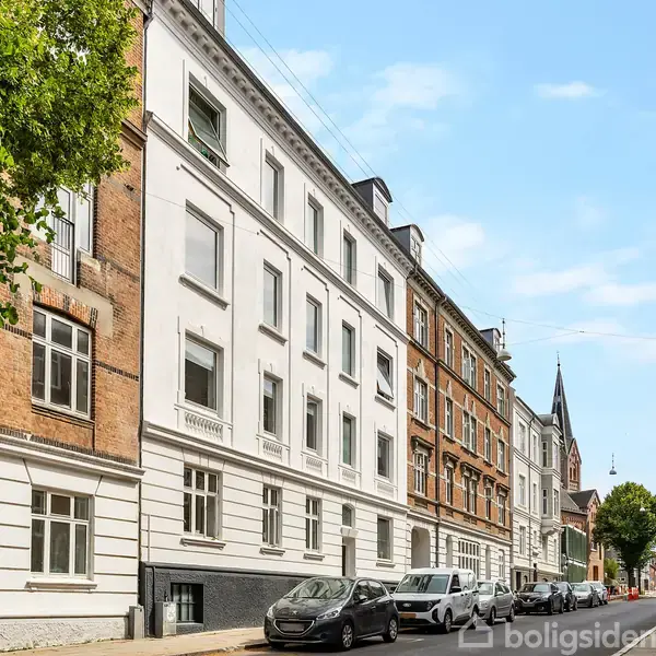 Bygning strækker sig langs en stille gade med parkerede biler foran. Bygningens facade veksler mellem hvidt og rødt murværk. Et handicapparkeringsskilt er synligt. Træ og kirketårn i baggrunden.
