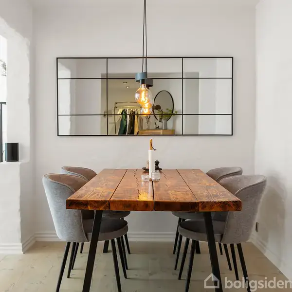 En rustik træspisebord står med fire grå polstrede stole. Over bordet hænger en glødepære fra en moderne lampe. En stor spejl vægreflekterer rummet. Tændt lysestage på bordet.