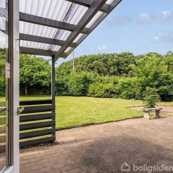 Terrasse med overdækning i bølgeplast står stille ved siden af flisebelagt havegang, omkranset af frodigt grønt græs og træer, under en blå himmel med få skyer.
