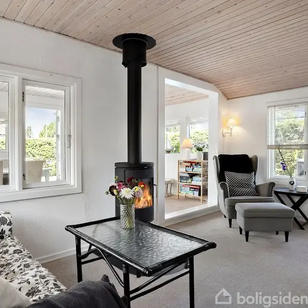 En moderne stue med en brændeovn centralt placeret, omgivet af hvide vægge og store vinduer. En sort lænestol står ved siden af et lille bord i et lyst rum.