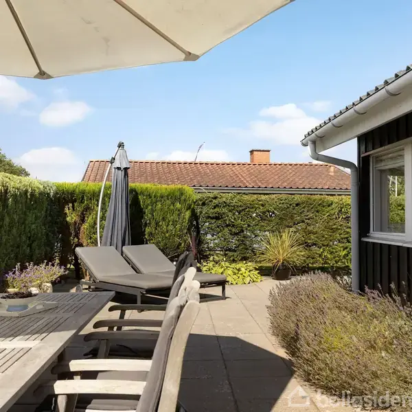 En terrasse med træmøbler og solsenge i solen. En stor parasol giver skygge. Omgivelserne inkluderer en hæk og et hus med rødt tegltag.
