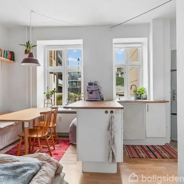 Et lyst køkken-alrum med træbord og stole, bogreol og hængende lampe. Køkkenet har hvide skabe, planter og kaffemaskine på bordpladen. Store vinduer giver udsyn til en bygning udenfor.