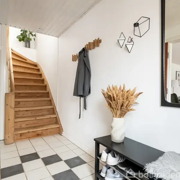 Trætrappe fører opad, flankeret af hvidmalede vægge. Væghængt jakke på knagerække; brun vase med tørrede planter står på sort bord. Fliser i sort/hvid mønster på gulvet.