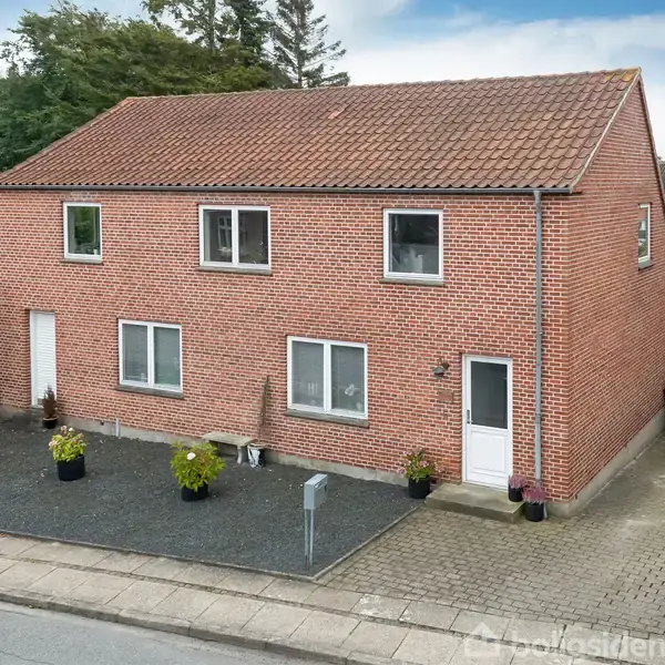 Rødt murstenshus med to etager står stille i et beboelsesområde. Foran huset er der en gårdsplads med potteplanter og en flisebelagt indkørsel.