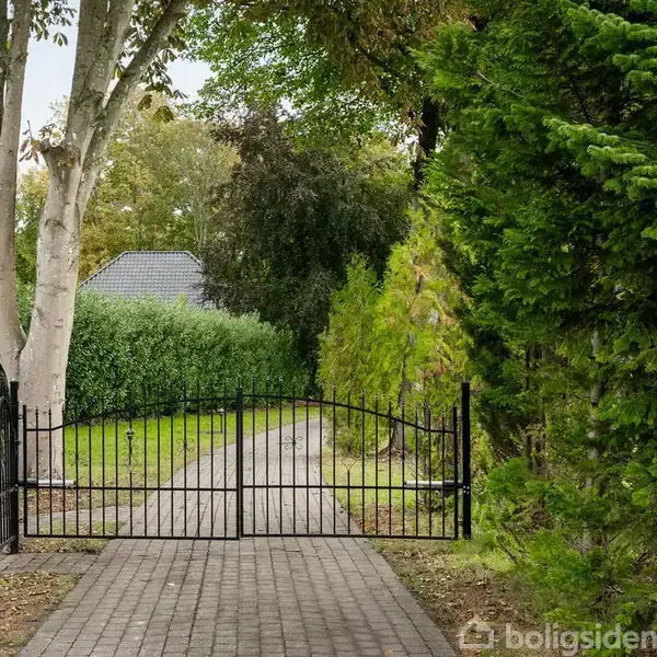 En sort metalport står åben på en brolagt sti omgivet af tæt, grøn bevoksning og træer, der fører til en have med et hus i baggrunden.