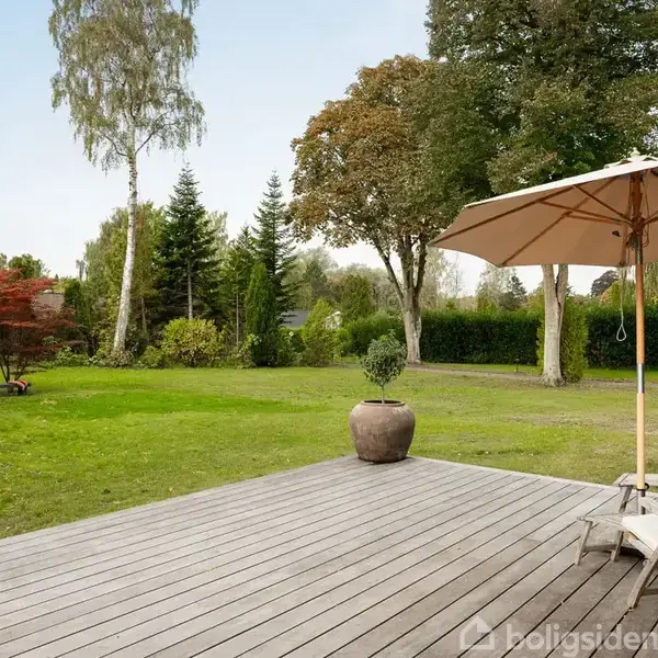 Terrasse med træmøbler og parasol står på trædæk, omgivet af frodig have med græsplæne og forskellige træer.