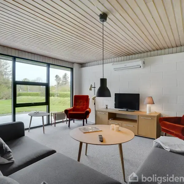 Stue med grå sofa og runde sofaborde, rød lænestol, fladskærms-tv på træskænk. Store vinduer med udsigt til grøn have. Træloft og hængende lampe.