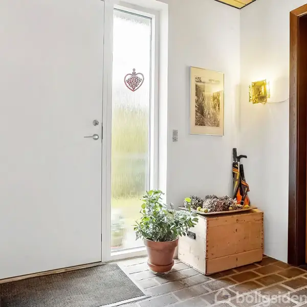 Dør i en entré står lukket, med et vindue i siden. En plante på gulvet, billeder på væggen og en træboks med paraplyer. Et gulvtæppe ligger foran døren.