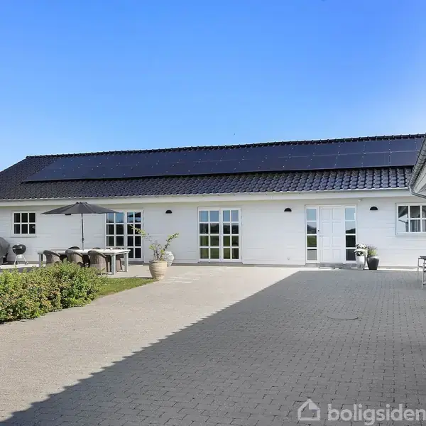 Et hvidt, moderne hus med solpaneler på taget står på en flisebelagt gårdsplads. Til venstre ses udemøbler og en have med grønne planter under en blå himmel.