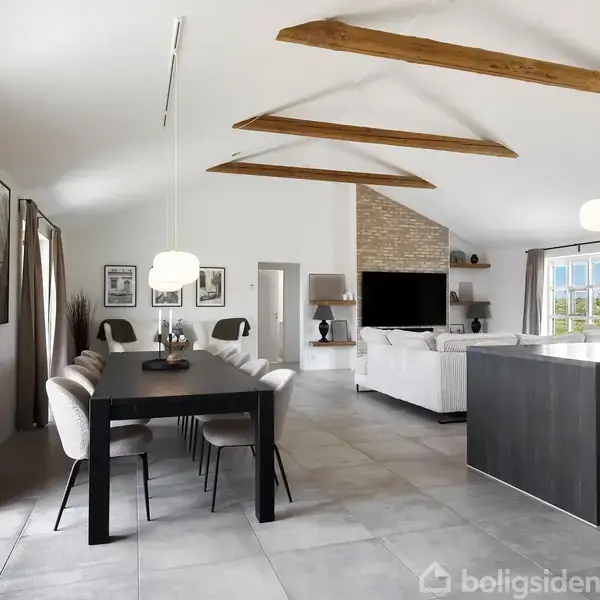 Stort, lyst opholdsrum med spisebord og stole til venstre. Køkkenø med barstole til højre. Højt loft med synlige bjælker. Store vinduer og pejs skaber en åben atmosfære.