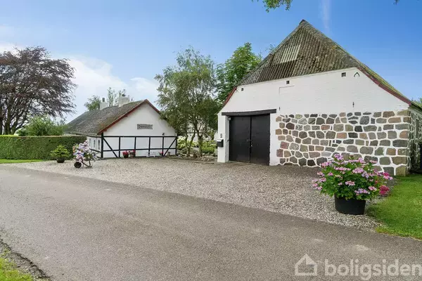 To huse med stenmure og sort/hvid bindingsværk står på en gårdsplads omgivet af grus, blomster og en landevej. Baggrunden består af træer og en blå himmel.