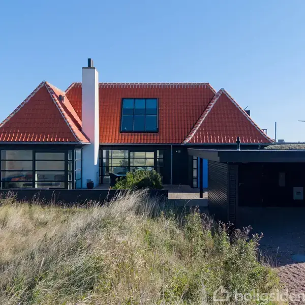 Et rødt teglhustag oven på et moderne hus med hvide vægge og store vinduer; en garage står til højre. Græsarealer omgiver huset, og der ses vindmøller i baggrunden.