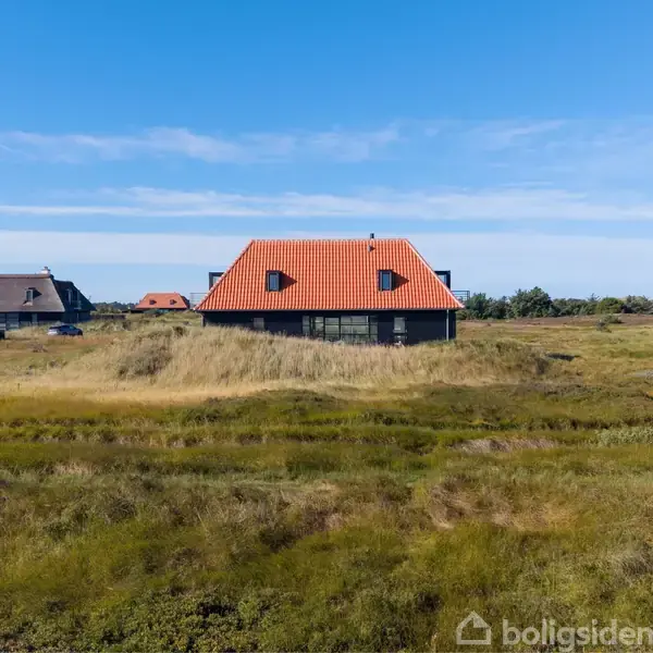 Et hus med rødt tag står stille blandt græsklædte klitter. Det er omgivet af flere huse og en blå himmel i baggrunden, hvilket skaber en fredelig landskabsudsigt.