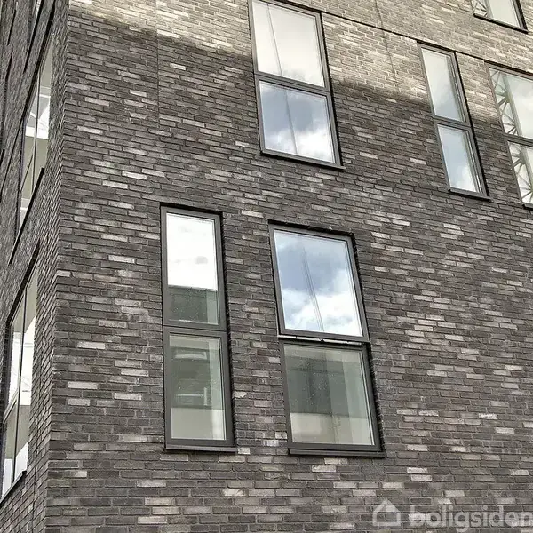 En murstensfacade med flere vinduer reflekterer himlen. Bygningen har en moderne arkitektur og tilstødende altaner. Det er dagtid, og himlen er delvist overskyet.