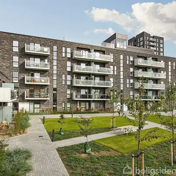 Et moderne lejlighedskompleks med altaner vender ud mod en grøn have med nyplantede træer og stier. Himlen er delvist overskyet, og bygningen er lavet af mørke mursten.