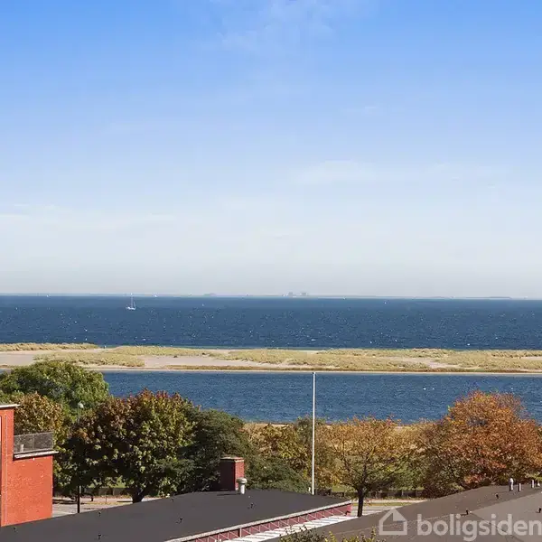 Et moderne boligbyggeri står stille ved kysten. Udsigten viser træer med efterårsfarver, en lang sandstrækning og blåt hav under en klar, blå himmel.