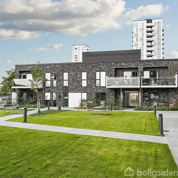 Moderne lejlighedsbygning med sorte mursten og store vinduer, omgivet af en velplejet græsplæne og stier. I baggrunden ses en højere bygning og himlen med få skyer.